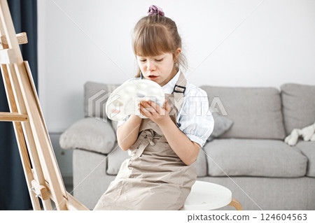 Little girl with Down syndrome wearing white shirt and beige apron. Girl is painting on easel. Girl sitting at home in living room. 124604563