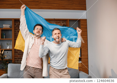 Male friends gathered at home to watch a football match. Two men standing and holding a big flag of Ukraine in hands. Men actively support their favorite team and feeling satisfied. 124604636