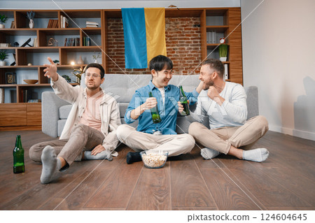 Male friends gathered at home to watch a football match. Men sitting on the couch in front of big flag of Ukraine on a wall. Men actively support their favorite team and feeling satisfied. 124604645