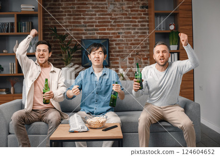 Front view of male friends gathered at home to watch a football match. Men sitting on the couch in front of the big screen TV and eating snacks. Men actively support their favorite team and feeling 124604652