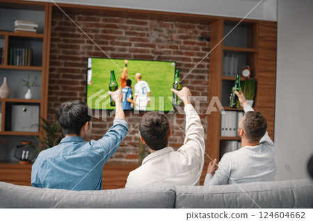 Back view of male friends gathered at home to watch a football match. Men sitting on the couch in front of the big screen TV. Men actively support their favorite team and comment on the match 124604662