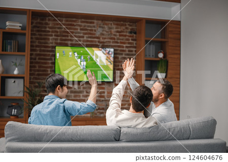 Back view of male friends gathered at home to watch a football match. Men sitting on the couch in front of the big screen TV. Men actively support their favorite team and comment on the match 124604676