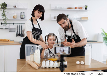 Caucasian family including adult man, woman, and child creating a cooking vlog in kitchen. Smiling while baking with whisk in bowl. Ingredients and camera focus on family moment. 124605600
