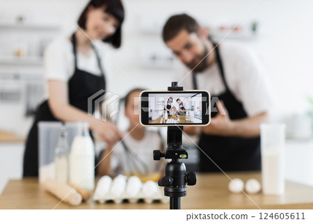 Caucasian family of blogger chefs working together kneading dough in glass bowl. Parents and child collaborating creatively on cooking a recipe while filming activity for social media on smartphone. Caucasian family of blogger chefs working together kneading dough in glass bowl. Parents and child collaborating creatively on cooking a recipe while filming activity for social media on smartphone. 124605611