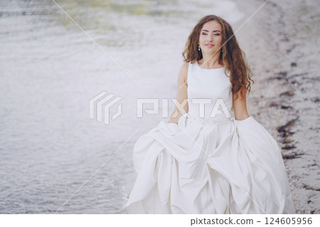 Beautiful long-haired bride in a magnificent white dress walking on a beach Beautiful long-haired bride in a magnificent white dress walking on a beach 124605956