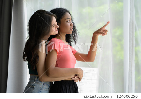 Happy diversity lesbian lgbtq couple hugging and standing next to the window at home in the morning. Happy diversity lesbian lgbtq couple hugging and standing next to the window at home in the morning. 124606256