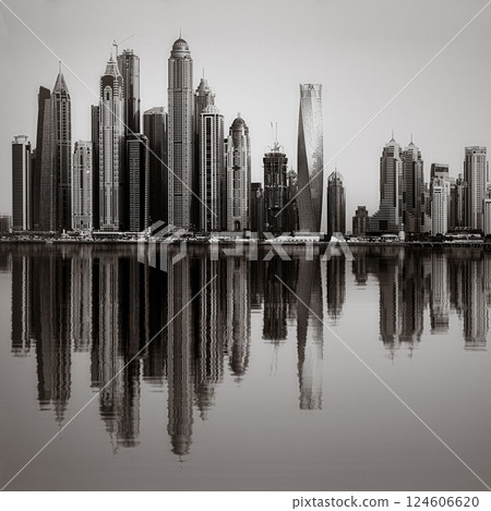 Monochrome city skyline with tall skyscrapers reflecting on calm water under a cloudy sky, Dubai Marina bay UAE 124606620