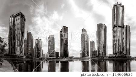 Monochrome city skyline with tall skyscrapers reflecting on calm water under a cloudy sky, Dubai Marina bay UAE 124606625