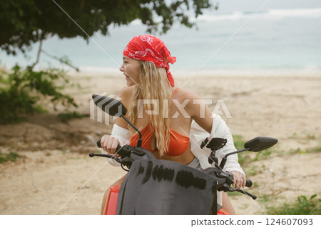 Attractive caucasian woman in a bikini swimsuit and on a bike scooter near the beach in sunny weather. Attractive caucasian woman in a bikini swimsuit and on a bike scooter near the beach in sunny weather. 124607093