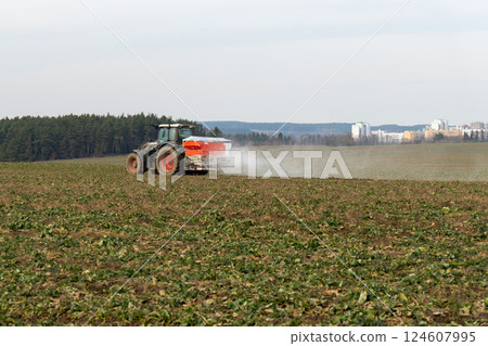 A tractor towing a mineral fertilizer dispenser across a green field 124607995