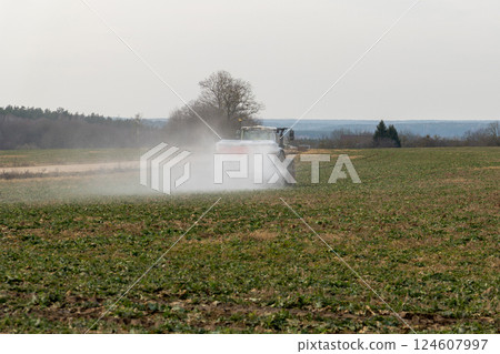 A tractor towing a mineral fertilizer dispenser across a green field 124607997