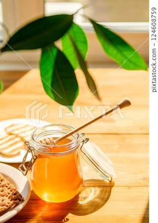 Honey Jar and Wooden Dipper on a Sunlit Table With Green Leaves and Cookies, Evoking a Warm Cafe Atmosphere Honey Jar and Wooden Dipper on a Sunlit Table With Green Leaves and Cookies, Evoking a Warm Cafe Atmosphere 124608759