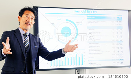 A middle-aged male business person giving an explanation in front of a large screen monitor 124610590