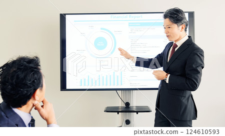 A middle-aged male business person giving an explanation in front of a large screen monitor 124610593