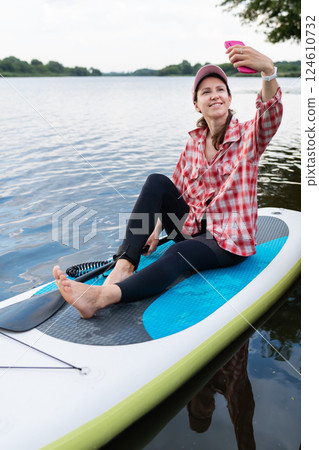A woman in a red plaid shirt and leggings sitting on a paddleboard, smiling while taking a selfie on a calm lake 124610732
