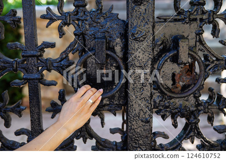 A woman's hand gently grasps the ornate ring pull of a dark, metal gate, showcasing intricate detailing and aged texture 124610752