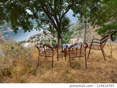Rustic metal chairs and a small table offer a scenic view of a bayside town nestled in a mountainous landscape 124610765