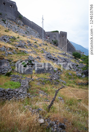 Ancient stone fortress walls climb a rocky hillside, showcasing weathered ruins and overgrown vegetation 124610766