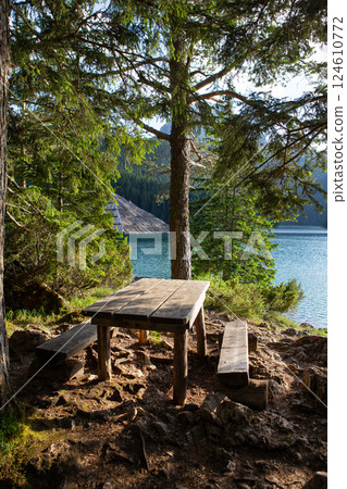 Rustic wooden picnic table and benches sit invitingly by a tranquil mountain lake, offering a peaceful lakeside retreat Rustic wooden picnic table and benches sit invitingly by a tranquil mountain lake, offering a peaceful lakeside retreat 124610772