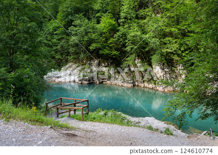 Serene turquoise river flows through a lush green forest, a wooden footbridge offers access to its tranquil banks 124610774