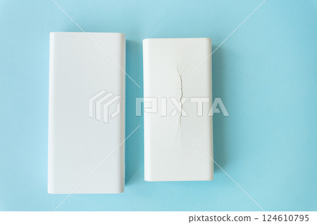 Two white rectangular power banks, one undamaged and the other cracked, rest on a light blue background 124610795