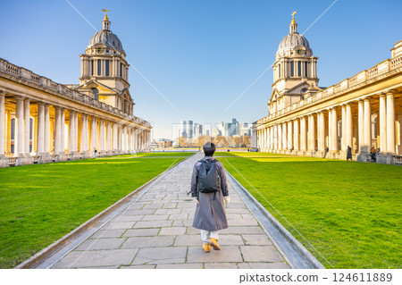 A visitor walks along the pathway amidst the stunning historical buildings of Greenwich Campus in London. With a clear blue sky, the city skyline can be spotted in the distance. A visitor walks along the pathway amidst the stunning historical buildings of Greenwich Campus in London. With a clear blue sky, the city skyline can be spotted in the distance. 124611889