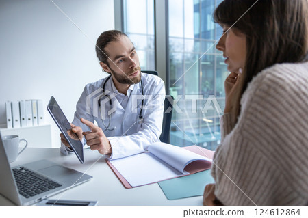 Male doctor sitting at desk in hospital holding tablet discussing digital health consultation Male doctor sitting at desk in hospital holding tablet discussing digital health consultation 124612864