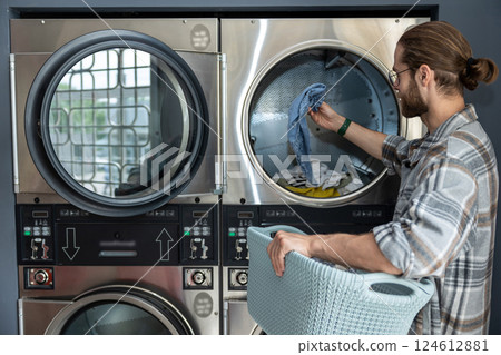 Man in public laundromat loading dirty clothes into washing machine 124612881