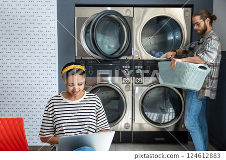 Woman working on laptop while bearded man doing laundry at public laundromat Woman working on laptop while bearded man doing laundry at public laundromat 124612883