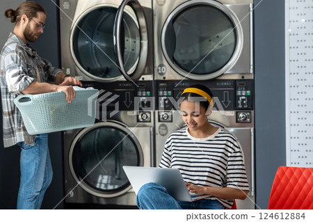Woman working on laptop while bearded man doing laundry at public laundromat Woman working on laptop while bearded man doing laundry at public laundromat 124612884