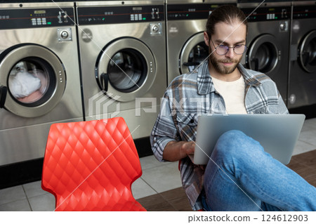 Man waiting for clothes to finish washing and using laptop for online work 124612903