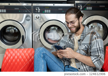 Bearded man in laundromat using washing machine while browsing online on mobile phone Bearded man in laundromat using washing machine while browsing online on mobile phone 124612906