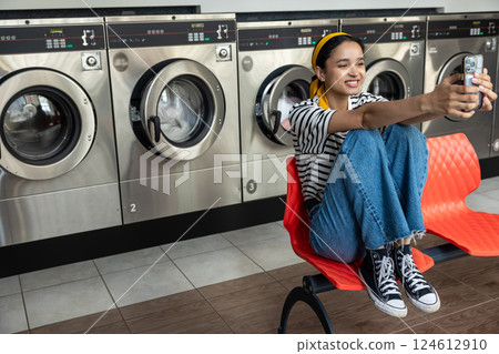Housewife in laundromat taking selfie on mobile phone while using washing machine 124612910