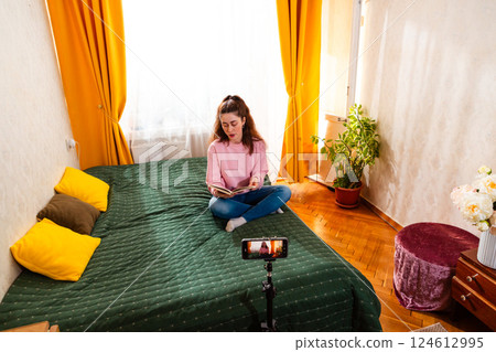 Caucasian woman sitting on a bed and creating a vlog using a smartphone and tripod, focusing on social media and online business strategies. Wide shot Caucasian woman sitting on a bed and creating a vlog using a smartphone and tripod, focusing on social media and online business strategies. Wide shot 124612995