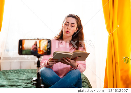 Caucasian woman sitting on a bed and making a video for her social media channel, using a tripod-mounted smartphone to communicate with her audience. Mid shot 124612997