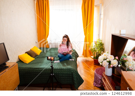 Young female entrepreneur recording a video on a smartphone tripod while sitting on her bed, engaging her audience in a vlog. Wide shot 124612998