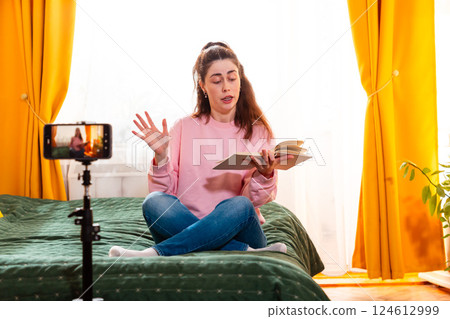 Young Caucasian woman filming herself at home while sitting on a bed, creating digital content for her social media presence. Mid shot 124612999