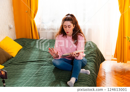 Female digital creator sitting on a cozy bed, recording a vlog with a smartphone on a tripod, sharing online business tips. Mid shot 124613001