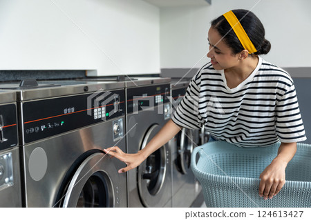 Woman in laundromat using self-service washing machine to clean dirty clothes in laundry store Woman in laundromat using self-service washing machine to clean dirty clothes in laundry store 124613427