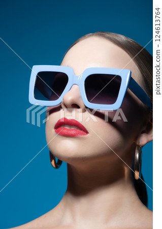 Woman portrait with classic hairstyle, big retro sunglasses, red lipstick and golden round earrings in blue background Woman portrait with classic hairstyle, big retro sunglasses, red lipstick and golden round earrings in blue background 124613764