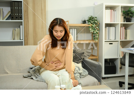 Asian woman suffering from stomach pain while sitting on sofa. Period cramps or gastrointestinal issues 124613849
