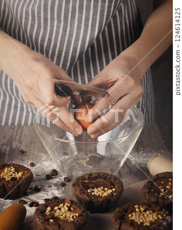 Human hands break eggs into a bowl next to muffins on on a table, faceless baker in striped apron, vertical image. Human hands break eggs into a bowl next to muffins on on a table, faceless baker in striped apron, vertical image. 124614125