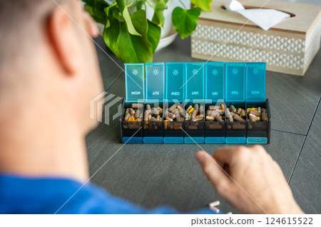Tired man looking at a full weekly pill organizer with medications and supplements. Concept of pill fatigue and overwhelming health routine 124615522