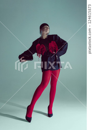 Confident young woman in black blazer, red tights, holding small handbag, striking angular pose against studio background. 124615873