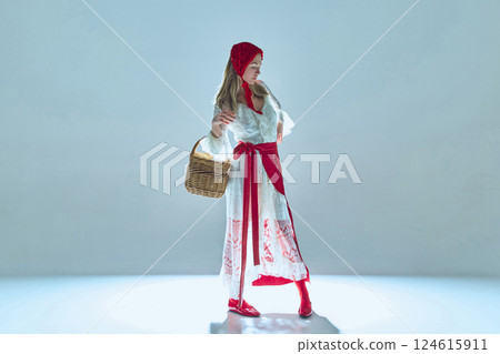 Young woman in white lace dress with red accents, evoking fairytale imagery, gracefully posing with basket, symbolizing contrast between innocence and power. 124615911