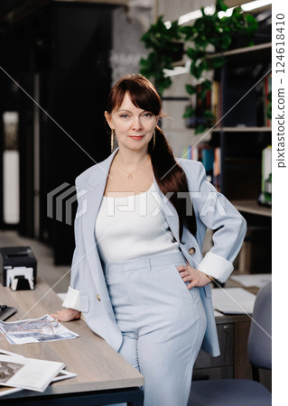 Businesswoman in a light blue suit standing near a desk with documents 124618410