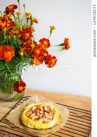 A homemade apple galette on a cooling rack accompanied by a lush marigold bouquet in a vase, set against a minimalistic backdrop A homemade apple galette on a cooling rack accompanied by a lush marigold bouquet in a vase, set against a minimalistic backdrop 124619215