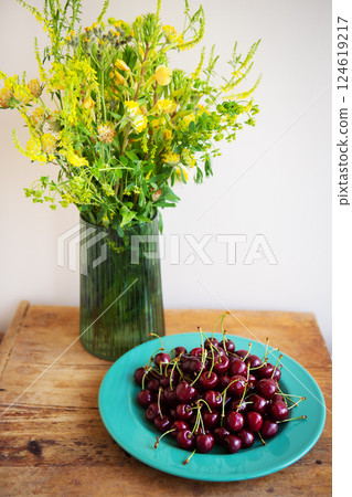 A vibrant summer still life featuring a bouquet of yellow wildflowers and a bowl of ripe red cherries on a rustic wooden table 124619217