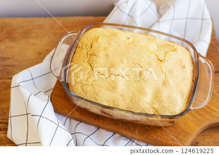 A golden-brown baked dessert, with a slightly cracked top, sits in a glass baking dish on a wooden board 124619225