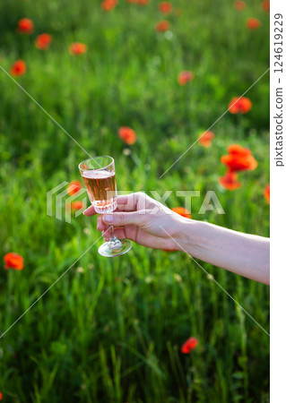 A hand holds a glass of rose wine against a blurred background of a vibrant green field with red poppies A hand holds a glass of rose wine against a blurred background of a vibrant green field with red poppies 124619229